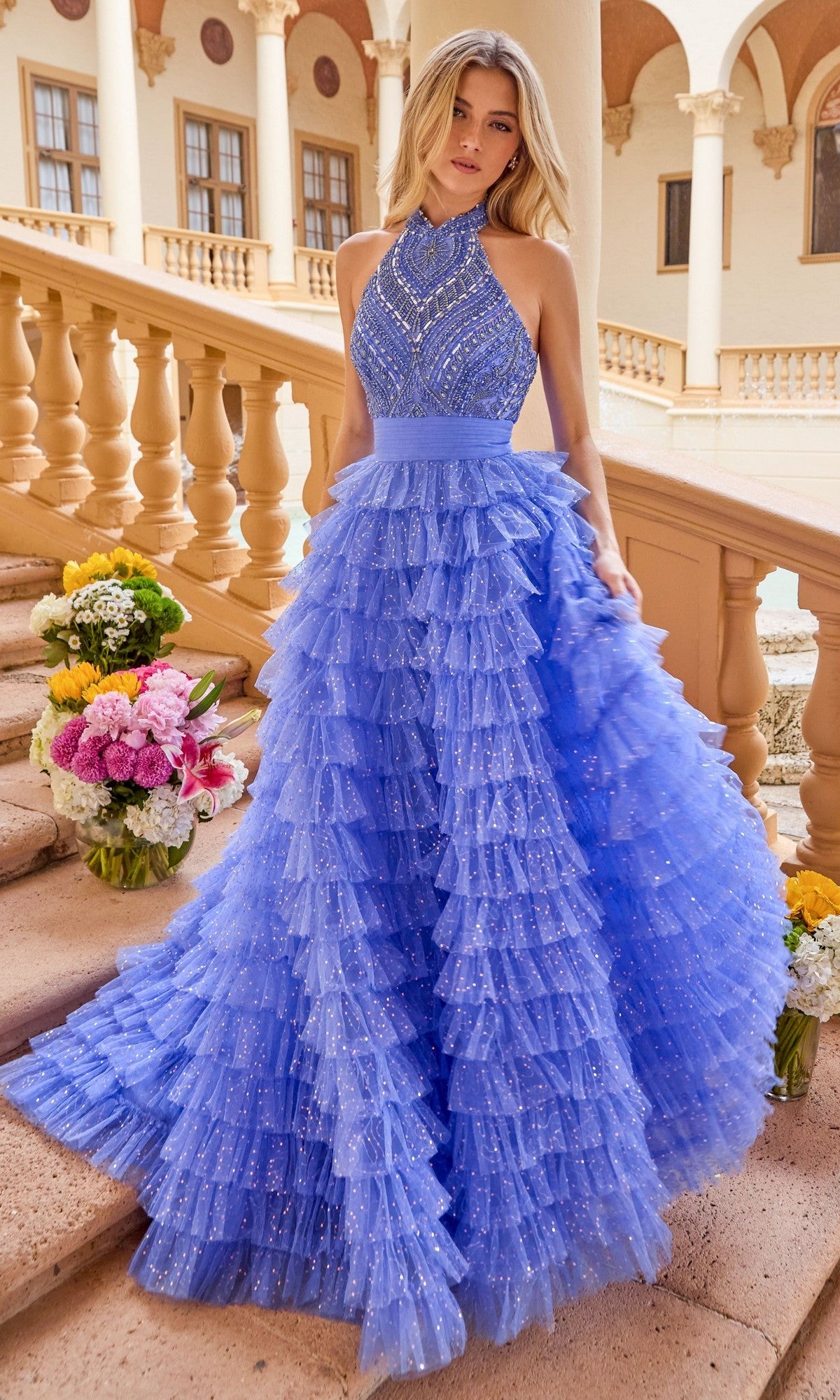 A woman stands on stairs wearing the Ava Presley 47258 long prom dress, a sparkling blue gown with ruffles and a tiered full skirt, bouquets of flowers nearby, and a columned building in the background.